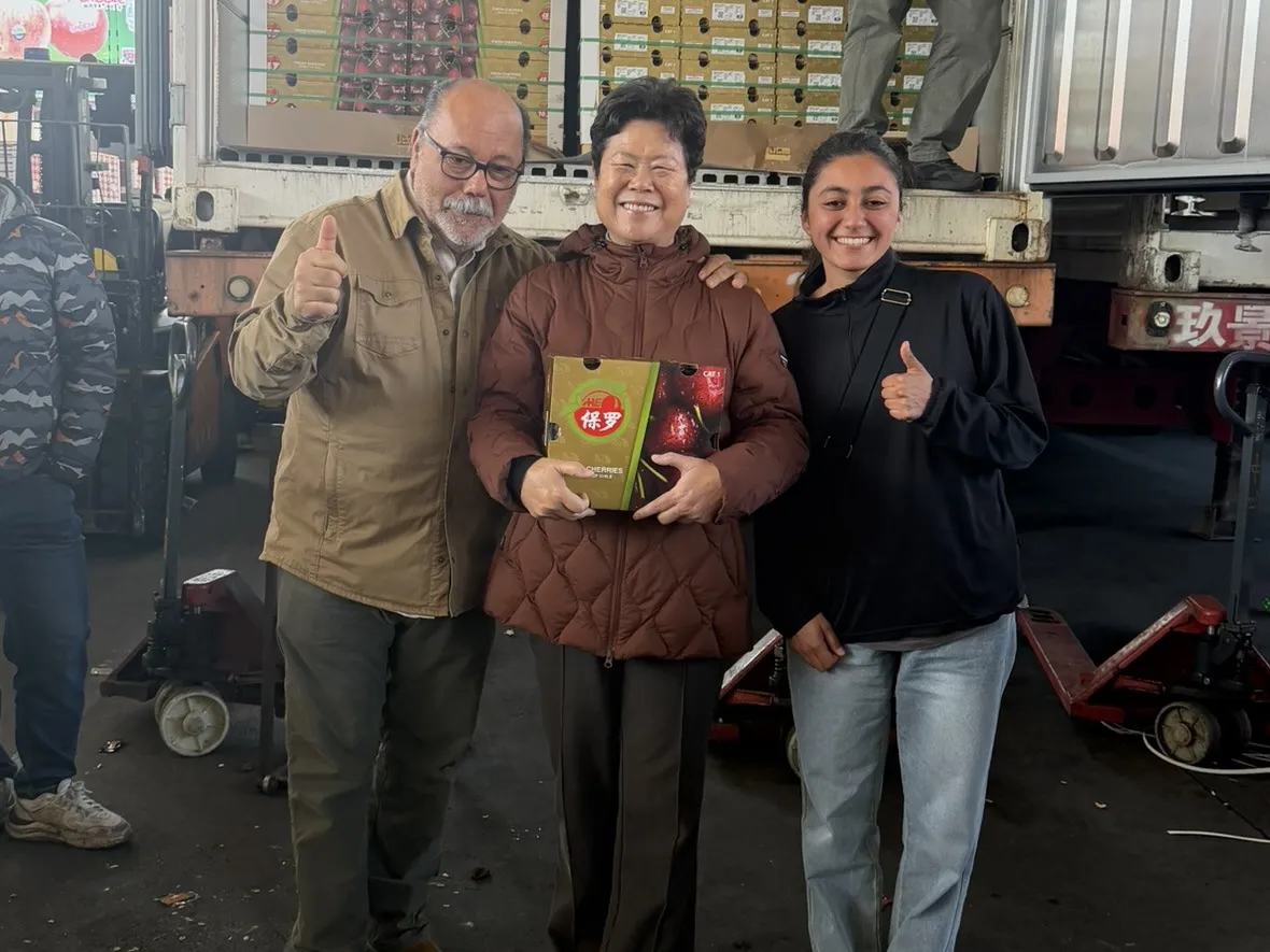 Un cliente sosteniendo una caja de cerezas, frente a un camión cargado de fruta para exportación
